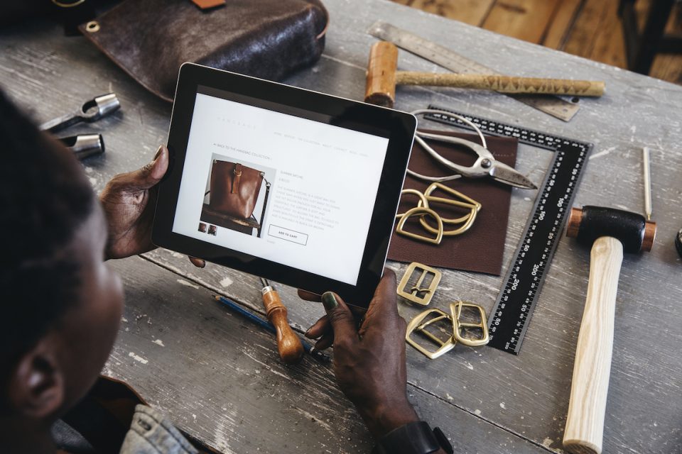 A leather craftswoman entrepreneur checks her website whilst sitting at her work bench. Leather goods and tools also lay on the desk.