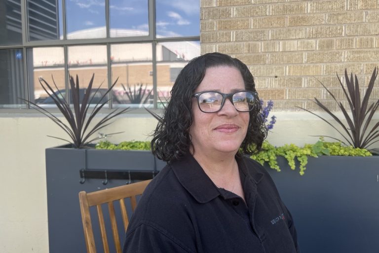 Latina woman in black polo shirt sitting outside the Senior Planet center in Colorado on a wooden chair