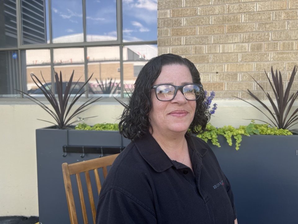 Latina woman in black polo shirt sitting outside the Senior Planet center in Colorado on a wooden chair