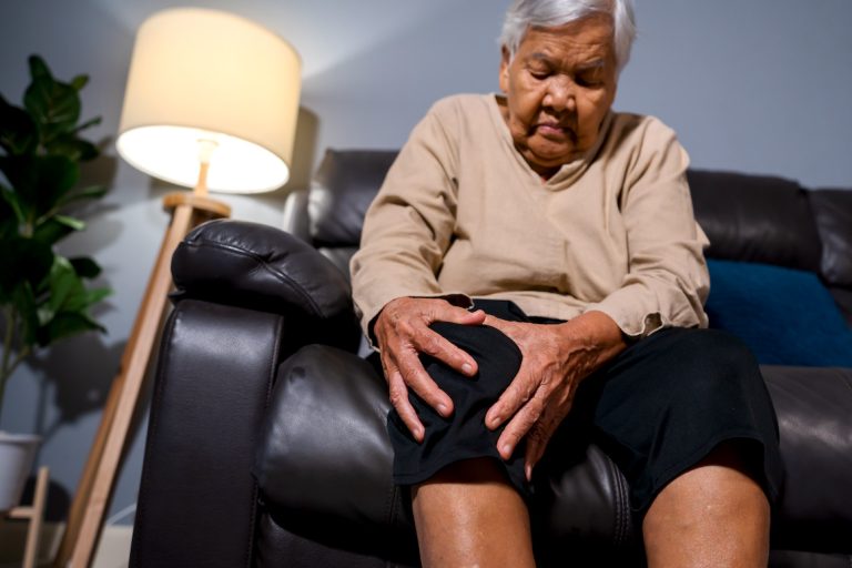 senior woman suffering from knee pain while sitting on a sofa in living room at night