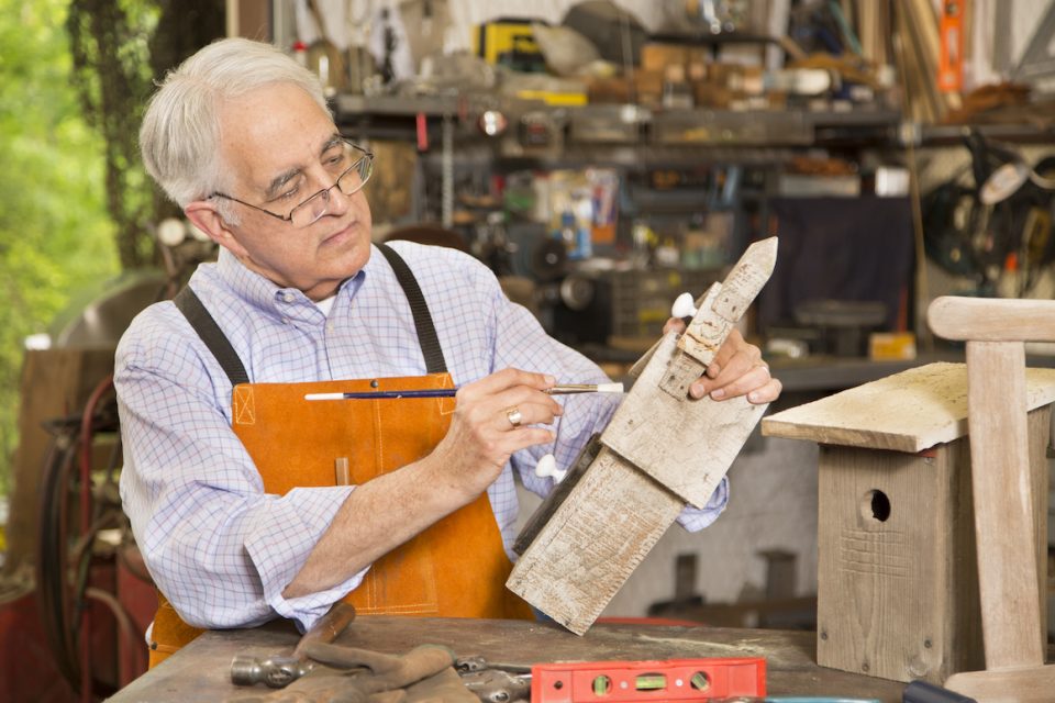 Senior man wearing orange apron works on refinishing two birdhouses in his workshop.