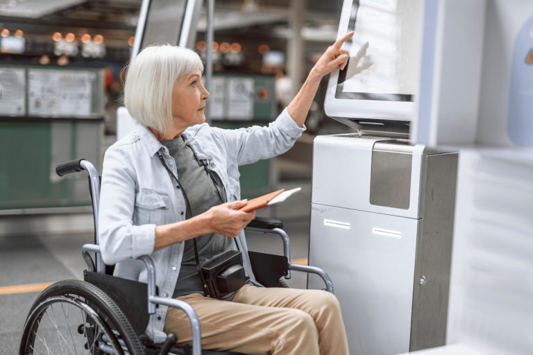 older woman with camera sitting in wheelchair and checking in at airport kiosk while holding ticket