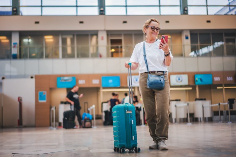 Cheerful elderly woman in glasses with a green suitcase in the lobby of the airport with a smartphone in her hands