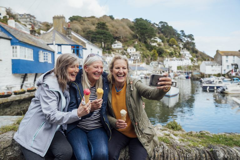 Three senior female friends sitting on a wall and taking a selfie together on a mobile phone while eating an ice cream in Polperro, Cornwall. The harbour is behind them.